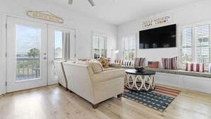 A living room with a beige sofa, a round coffee table, and a TV mounted on the wall. There are two doors, one labeled "Beach House" and another with a sign reading "Gulf Shores."