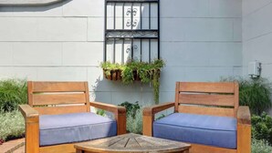 Dos sillas de madera con cojines azules y una pequeña mesa de madera frente a un edificio con pared blanca y una reja de metal.