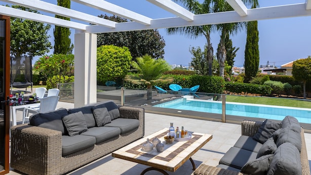 Un patio al aire libre con mobiliario de mimbre, piscina y vistas de la vegetación y edificios circundantes.
