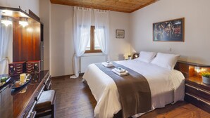 Un dormitorio con techo de madera, una cama con ropa blanca, una mesita de noche, un aparador y una ventana con cortinas traslúcidas.