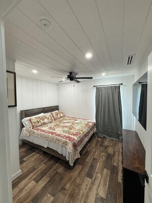 Quarto com piso de madeira, cama com colcha florida, ventilador de teto e janela com cortina.