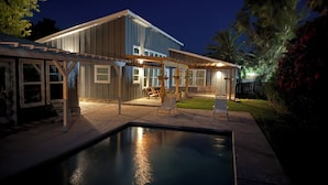A modern house with a covered patio, a swimming pool, and a well-lit garden at night.