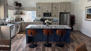 A modern kitchen with light wood cabinets, a central island with bar stools, stainless steel appliances, and a hanging light fixture.