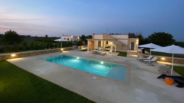 Una piscina rectangular con borde de piedra, rodeada por un patio empedrado. Hay sillones de descanso y sombrillas bajo una zona techada.