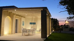 Un patio techado al aire libre con una mesa y sillas, un pilar de piedra y vistas al atardecer.