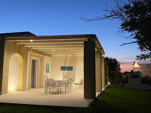 An outdoor covered patio with a dining table and chairs, a stone pillar, and a view of the sunset.