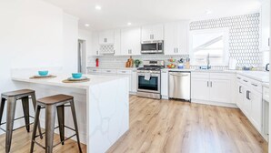 Cozinha moderna com armários brancos, uma ilha central com banquetas, micro-ondas, fogão e lava-louças.