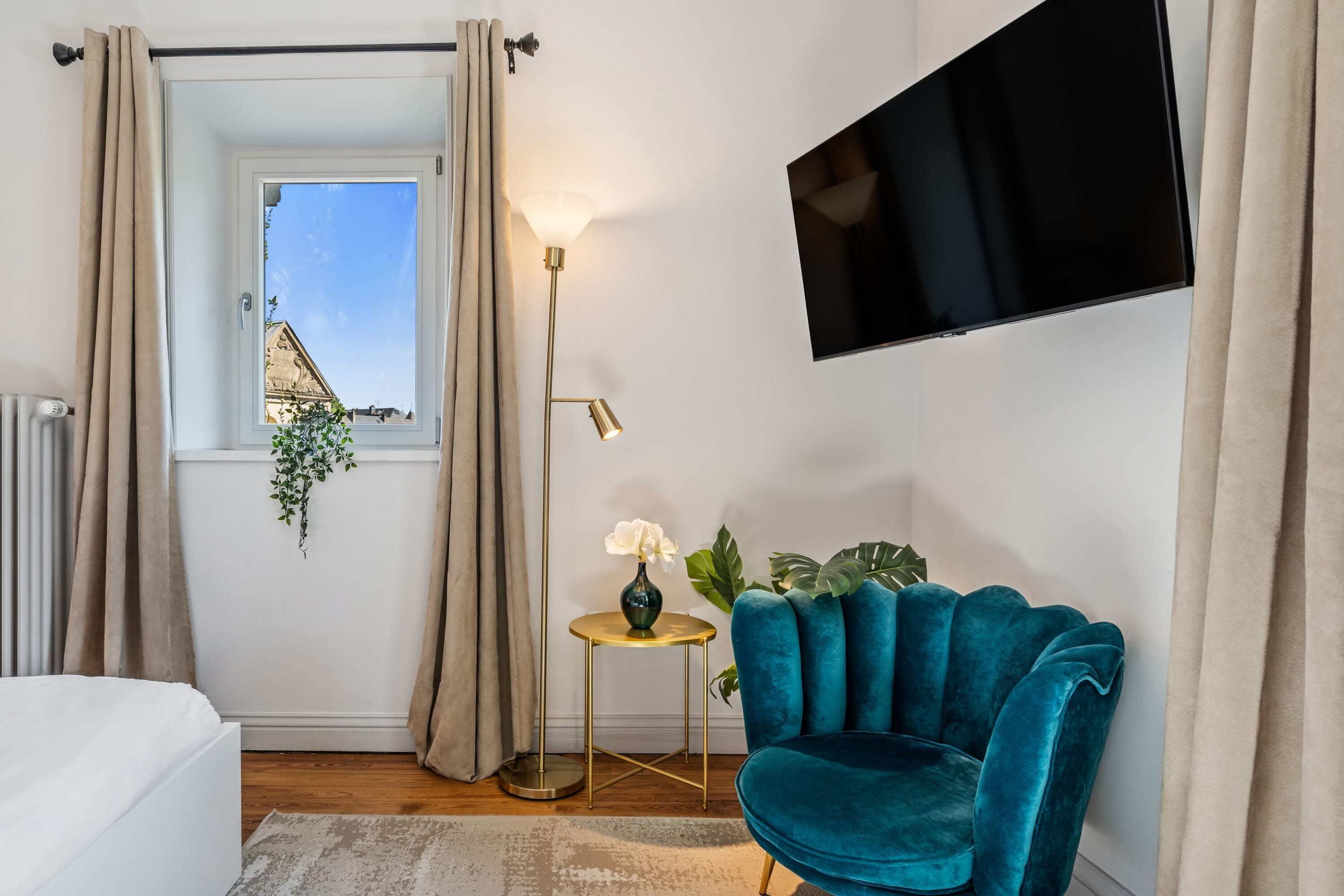 Et moderne soveværelse med en seng, en turkis lænestol, et lille rundt bord med en lampe, et fjernsyn monteret på væggen og et vindue med gardiner.