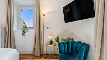 Et moderne soveværelse med en seng, en turkis lænestol, et lille rundt bord med en lampe, et fjernsyn monteret på væggen og et vindue med gardiner.