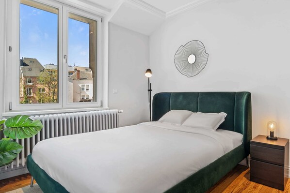 Un dormitorio con una cama grande, una lámpara de noche, un radiador y vistas a edificios.