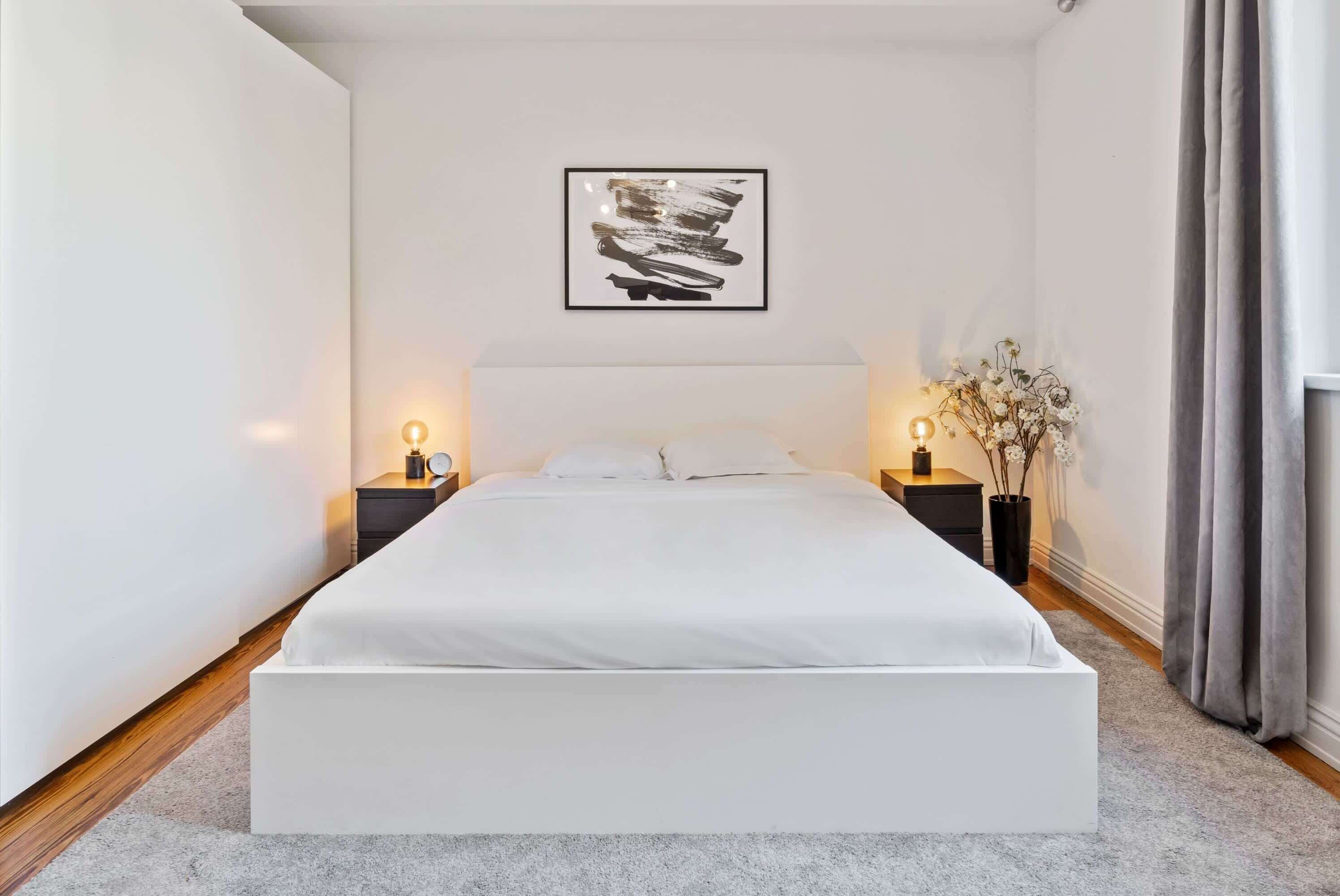 Ein modernes Schlafzimmer mit einem großen Bett, zwei Nachttischen mit Lampen, einem gerahmten abstrakten Kunstwerk an der Wand und einer Vase mit Blumen auf der rechten Seite.