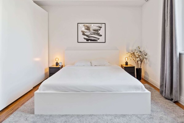 A modern bedroom with a large bed, two bedside tables with lamps, a framed abstract artwork on the wall, and a vase with flowers on the right side.