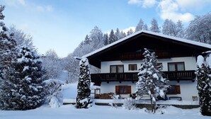 A two-story chalet with a wooden balcony, surrounded by snow-covered trees and a mountain backdrop. - Ferienwohnung 2 Personen, 40 qm, Jenner, Sep. Schlafzimmer, Balkon (Schönau am Königssee)