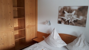 A bedroom with a wooden wardrobe, a bed with checkered pillows, a wall-mounted picture of a flower, and a bedside table with a lamp. - Ferienwohnung 2 Personen, 50 qm, - Hoher Göll, Sep. Schlafzimmer, Balkon (Schönau am Königssee)