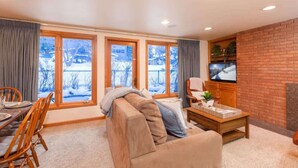 A cozy living room with a sofa, a coffee table, and a TV. There are large windows with a view of a snowy landscape. - Comfort & Location - 2 BR, 2BA - Walk to Gondola! (Aspen)