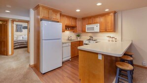 A compact kitchen with wooden cabinets, a white refrigerator, a microwave, a stove, and a small island with stools. - Comfort & Location - 2 BR, 2BA - Walk to Gondola! (Aspen)