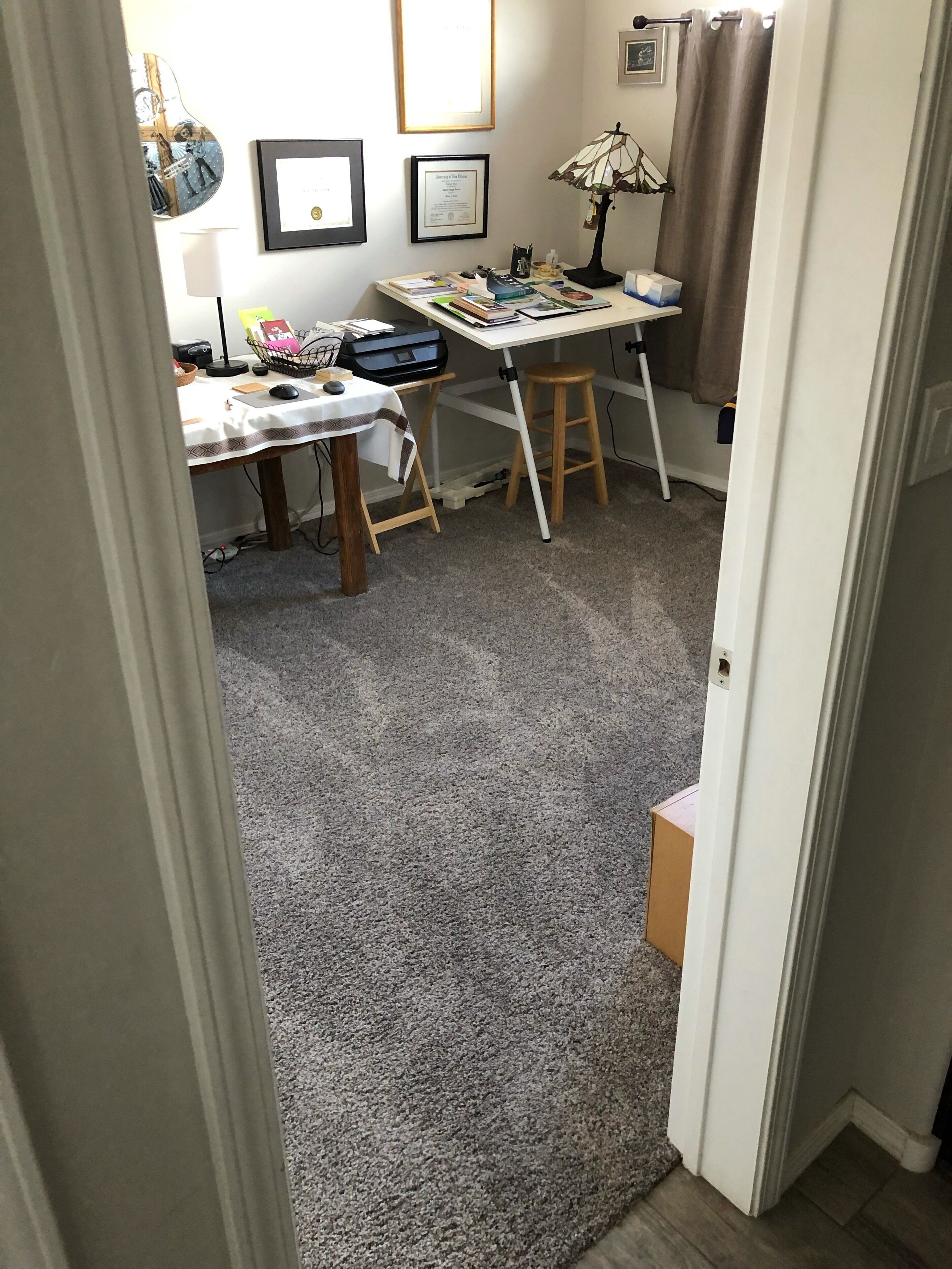 Una habitación con escritorio, silla y un taburete pequeño. En la pared hay diplomas enmarcados, una lámpara y una decoración con forma de guitarra.