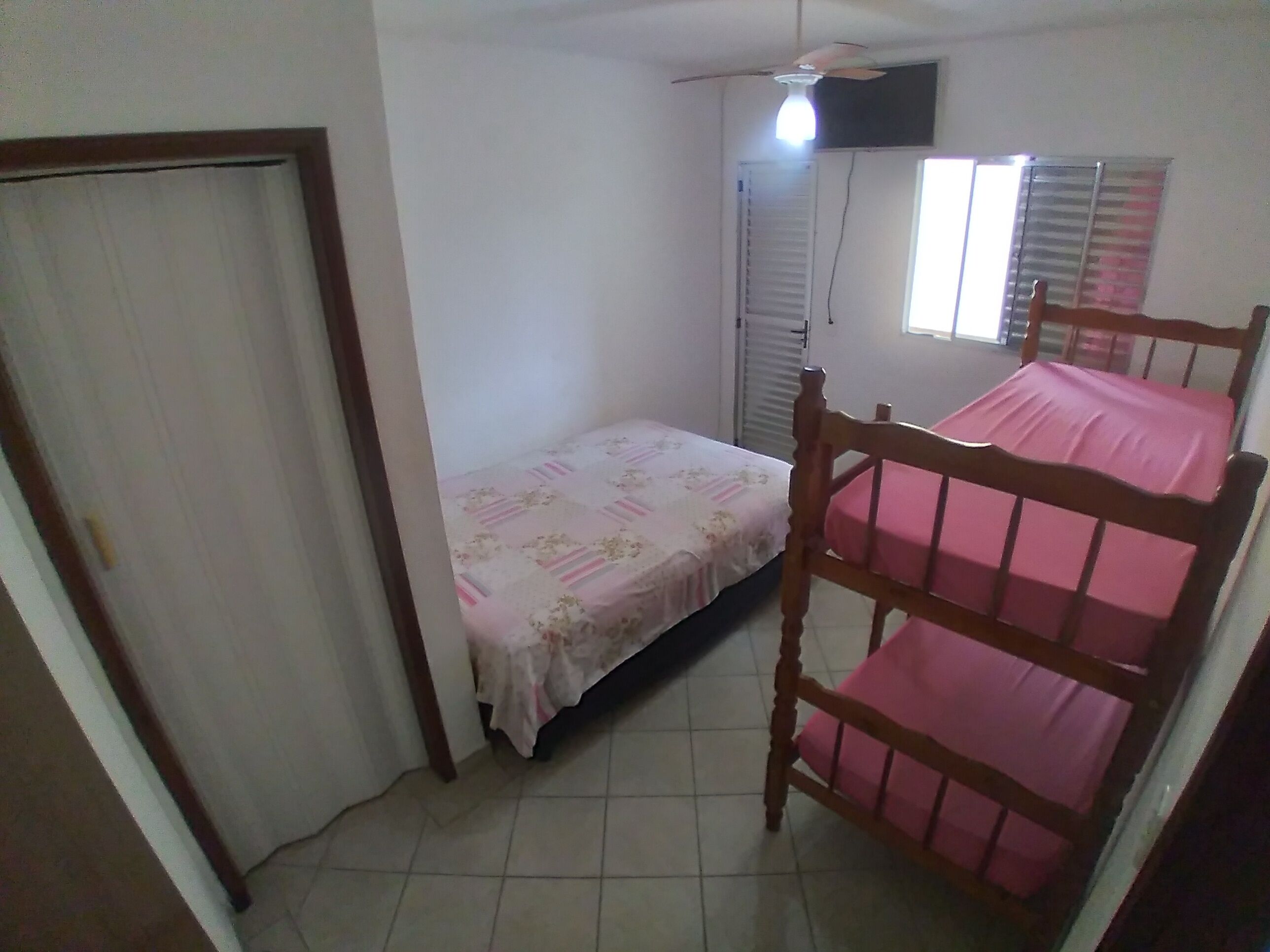 Een kleine, eenkamer accommodatie met een bed, een houten stoel, een plafondventilator, een raam met zonwering en een deur.