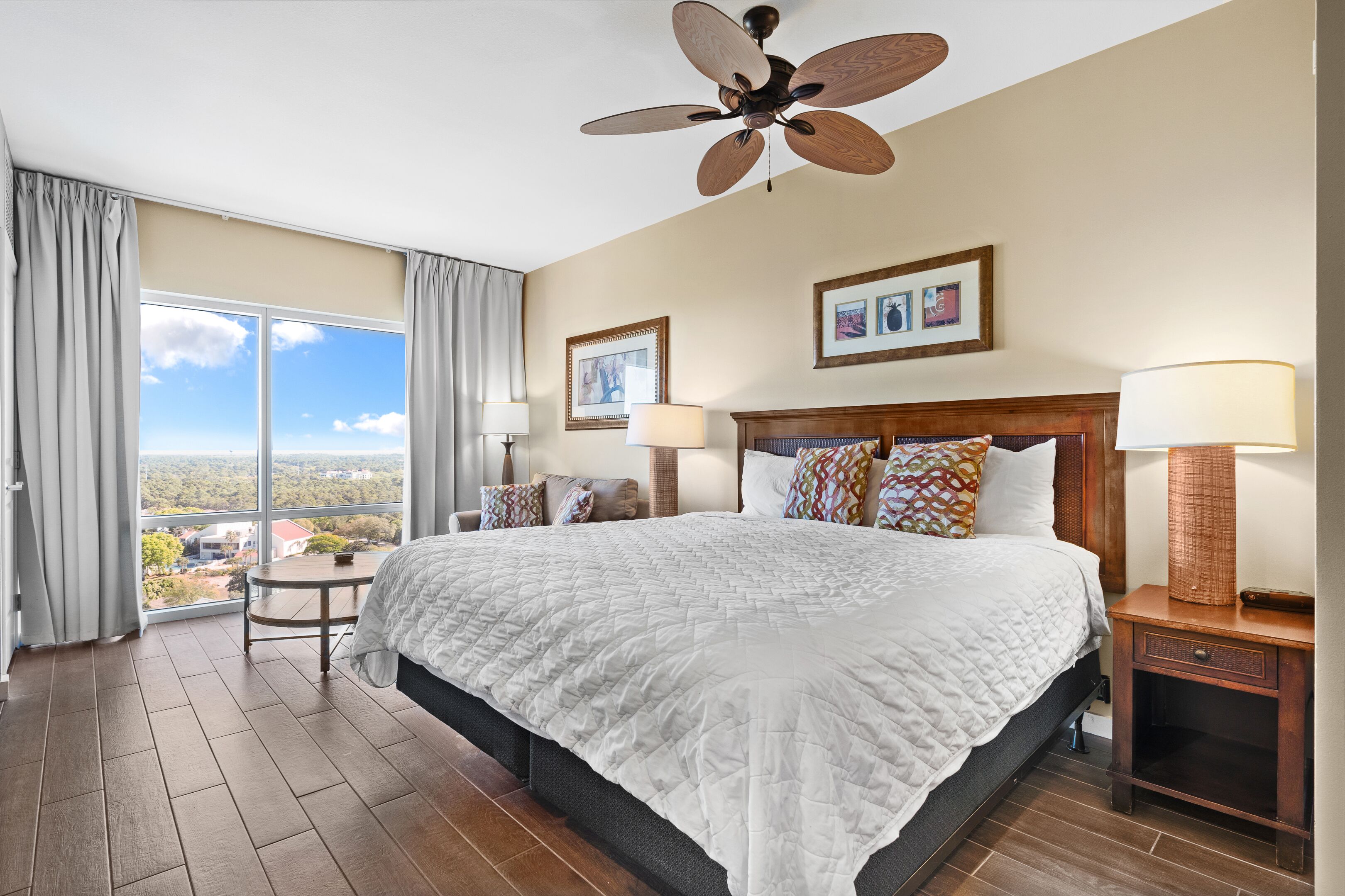 Ein Hotelzimmer mit einem großen Bett, einem Deckenventilator, Blick auf die Stadt und einem kleinen Tisch mit einer Lampe.