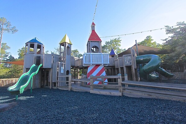 A wooden play structure with slides and climbing areas, surrounded by gravel.
