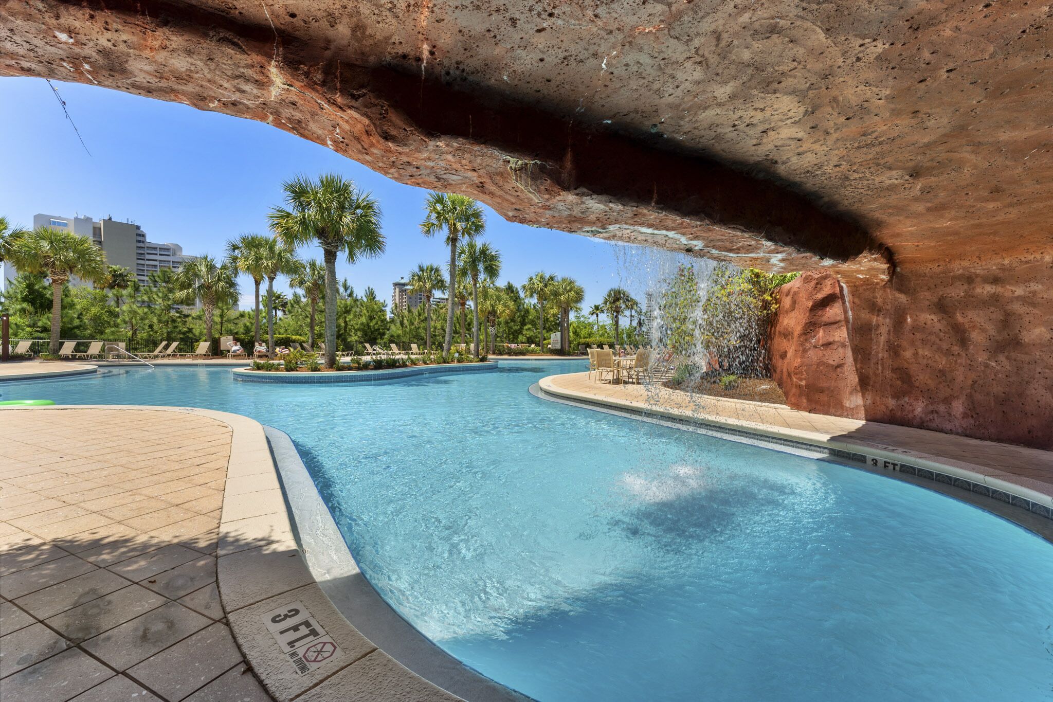 A swimming pool with a natural rock arch overhead, surrounded by palm trees and lounge chairs.