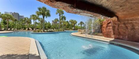 A swimming pool with a natural rock arch overhead, surrounded by palm trees and lounge chairs.