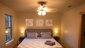 A bedroom with a large bed, two bedside tables with lamps, a ceiling fan, and three wall-mounted flower paintings.