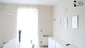 Une cuisine moderne et lumineuse, avec un plan de travail blanc, une table à manger et des chaises. Des tableaux encadrés ornent le mur, et un radiateur est placé en dessous.