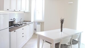Une cuisine moderne avec des meubles blancs, un four à micro-ondes encastré et une cuisinière à gaz. On y trouve une table à manger avec des chaises et un vase contenant des fleurs séchées.