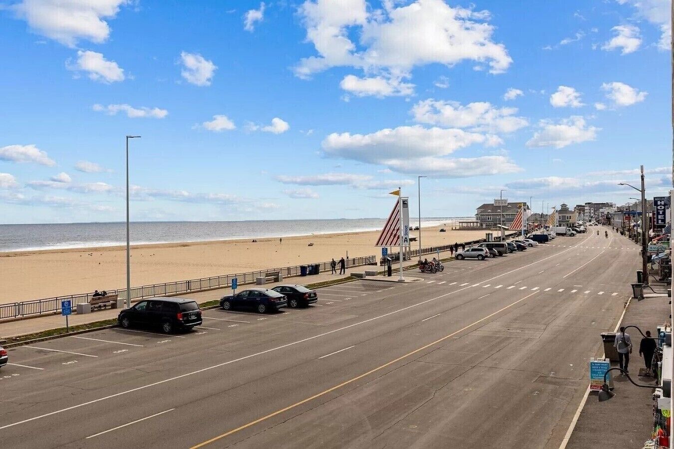 A beachside road with parked cars, a flag, and buildings including a hotel.