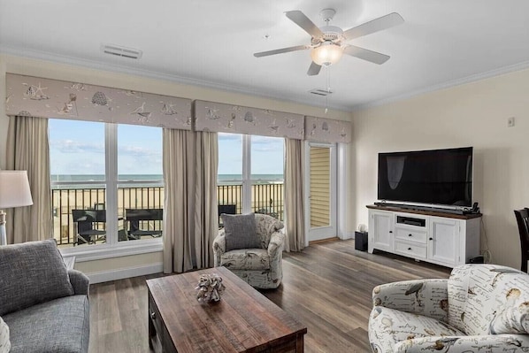A living room with a TV, a ceiling fan, and a view of the beach.