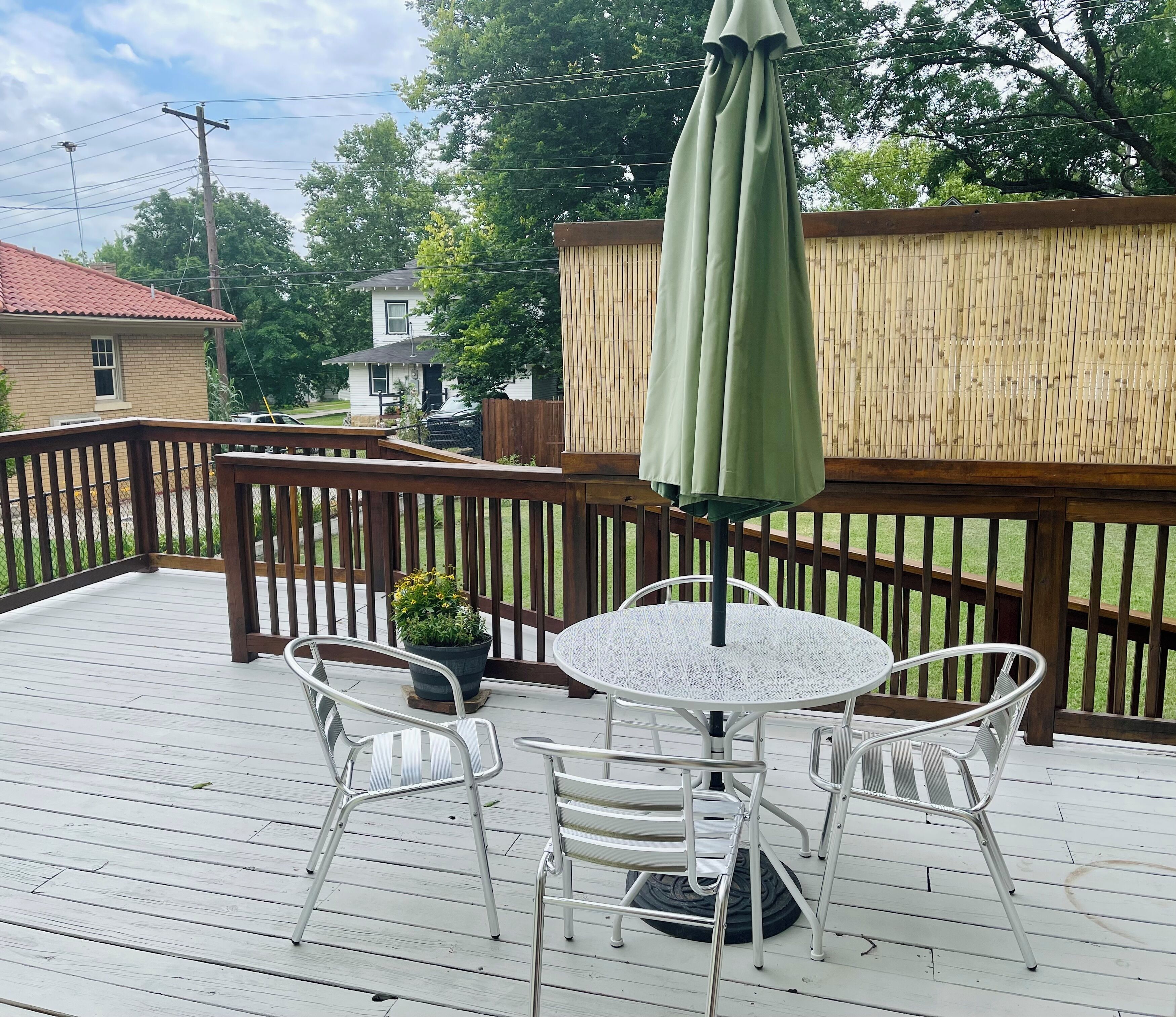 Een terras met een ronde tafel en twee stoelen, een groene parasol en een houten omheining.