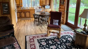 L’intérieur d’un chalet rustique, avec des murs en bois, un coin repas comprenant une table et des chaises, une cuisine équipée et une grande fenêtre donnant sur le paysage extérieur.