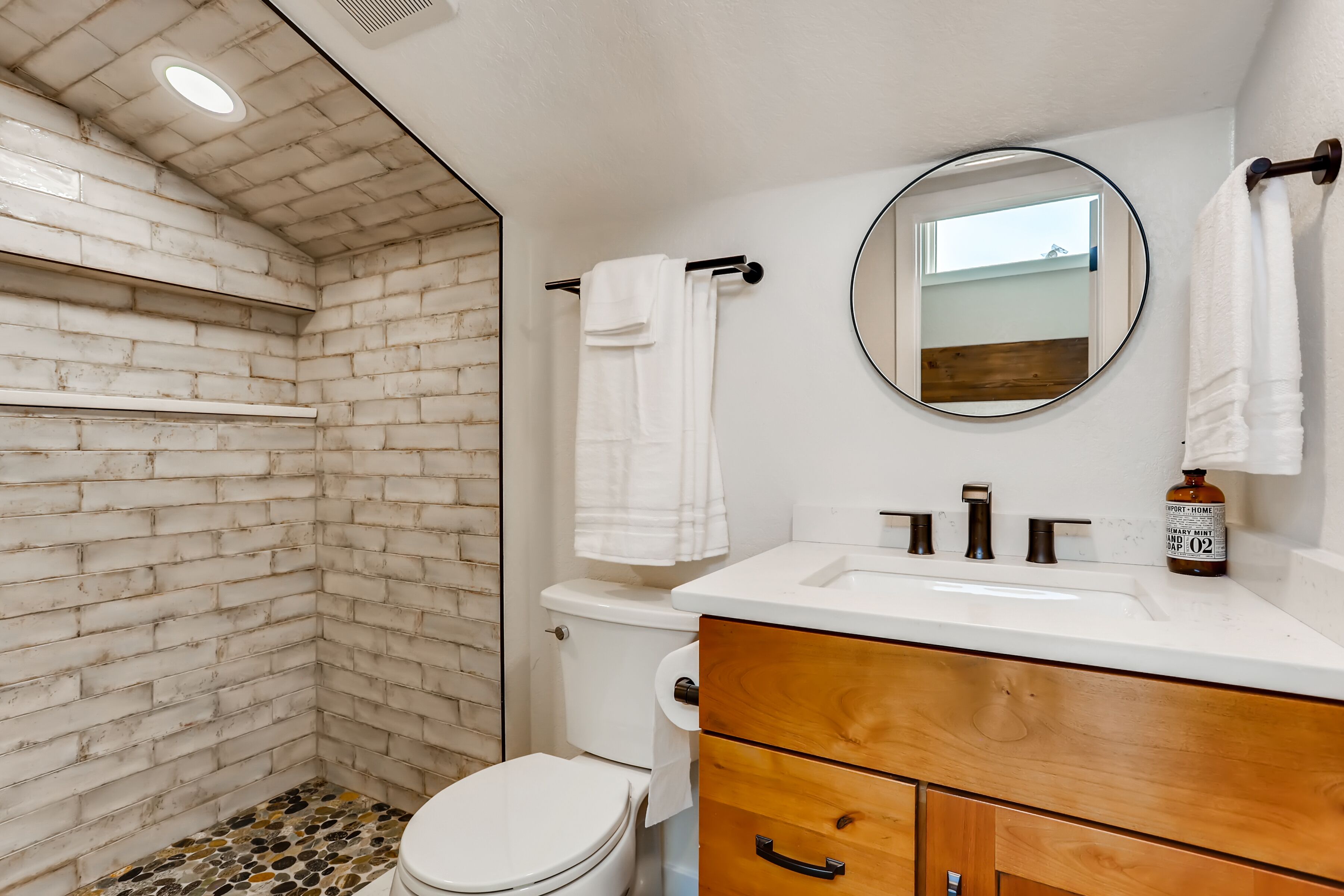 A bathroom with a white brick wall, a white toilet, a wooden vanity with a mirror, and a sink with two faucets.