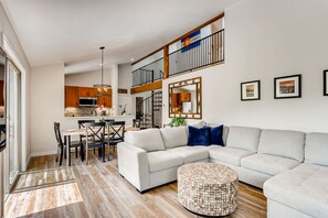 Een ruime woonkamer met een grote L-vormige zithoek, een rond hockertje en een eettafel met stoelen. Een trap leidt naar een verdieping met een balkon.