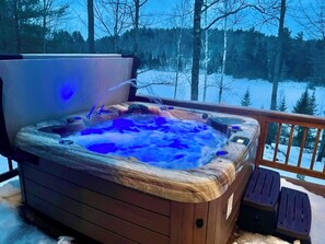 Un bain à remous avec une structure extérieure en bois, entouré d’une terrasse en bois, avec un paysage enneigé et des arbres en arrière-plan.