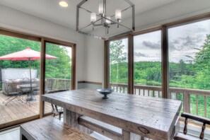 Une terrasse en bois avec une table et des chaises, un parasol rouge et une vue sur une forêt, visible à travers de grandes fenêtres.