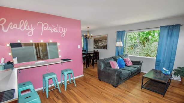 Una habitación con una pared rosa, un tocador con luces, un sofá gris y una ventana con cortinas.