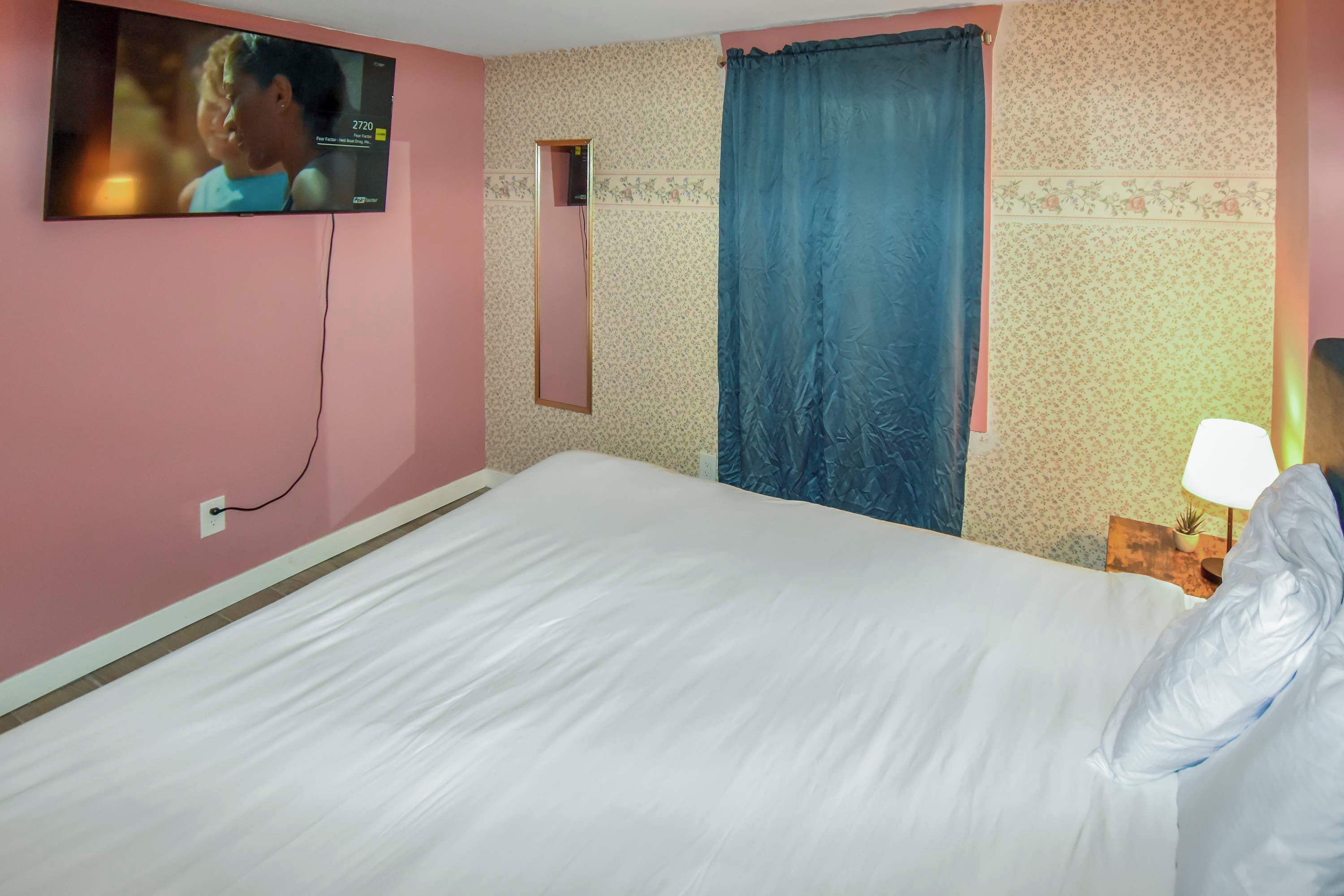 A hotel room with a bed, a television mounted on the wall, a mirror, a window with curtains, and a bedside table with a lamp.