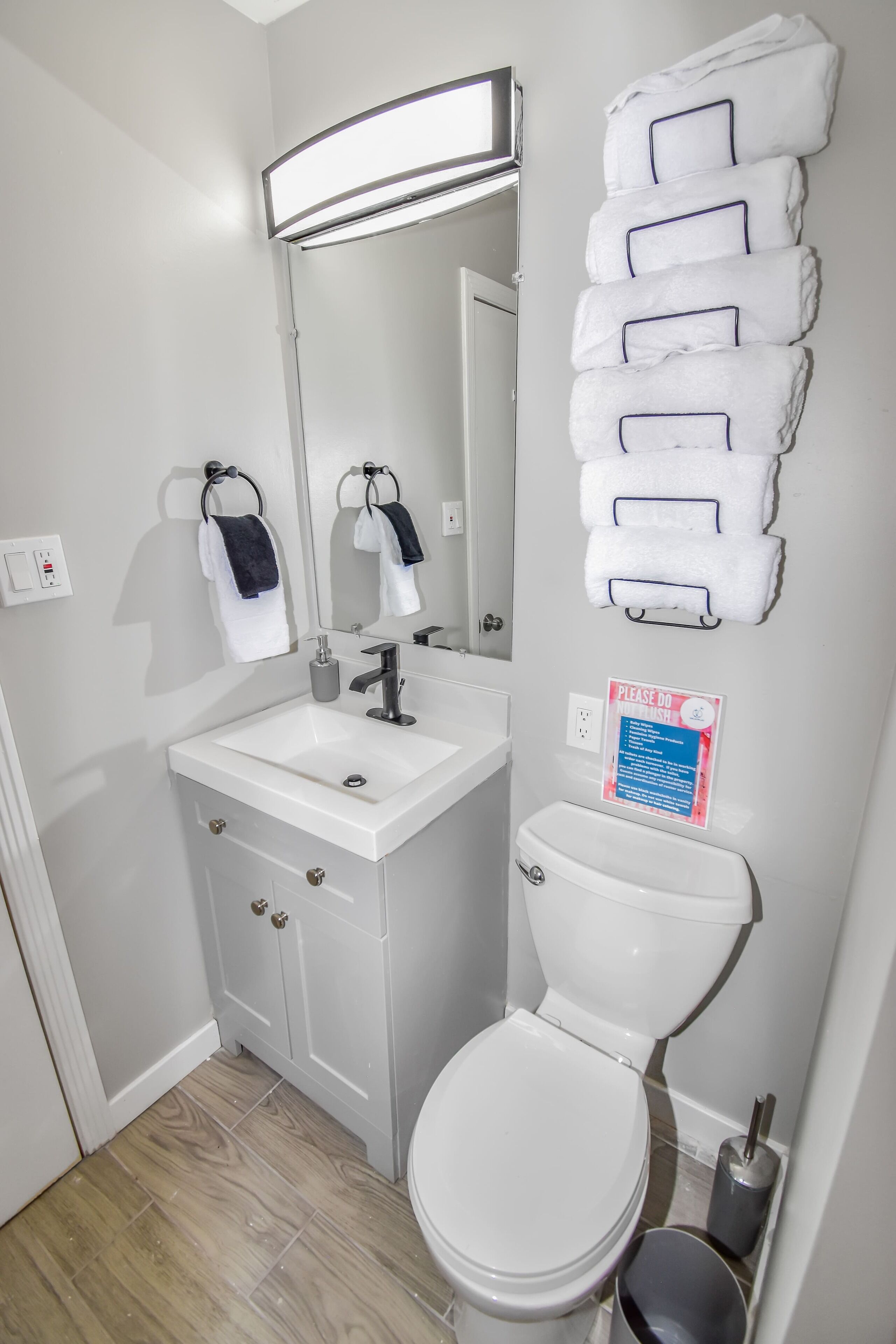 Een badkamer met een wit wastafel, spiegel en toilet. Handdoeken hangen aan een rek en er is een bordje aan de muur.