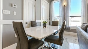 A modern dining area with a white table and four chairs, a potted plant, and a view of the city through large windows.