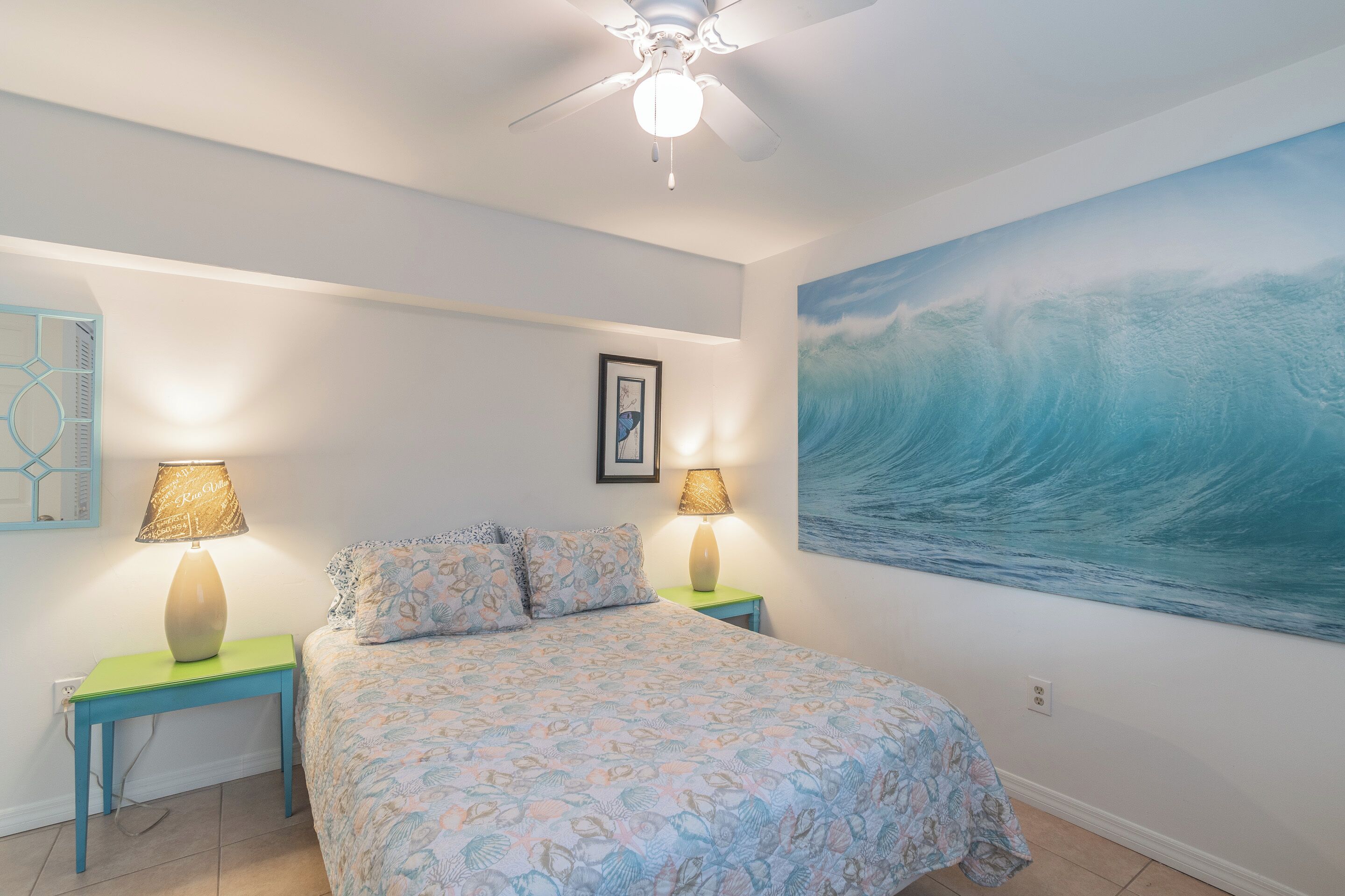 Une chambre à coucher comprenant un lit, deux tables de chevet, un ventilateur de plafond, un tableau mural et un tableau encadré.