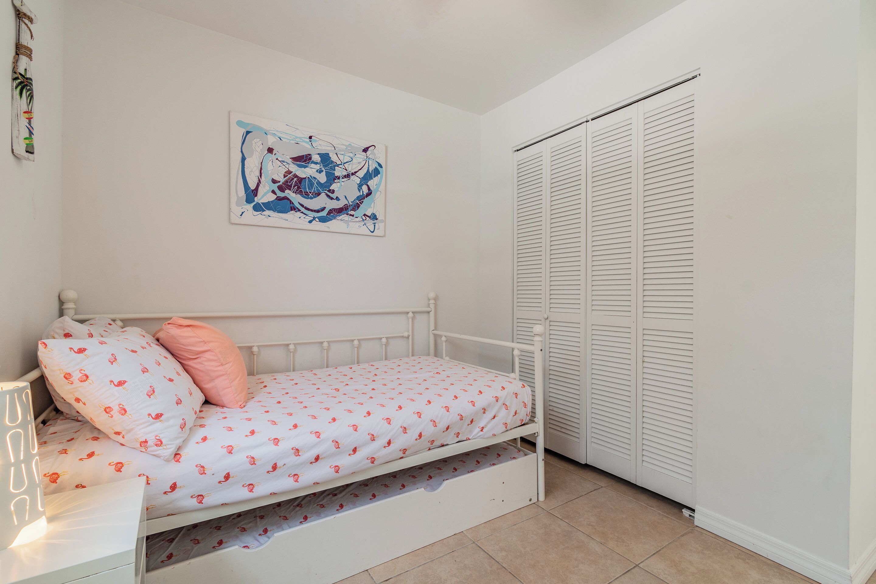 Uma cama branca com travesseiro rosa e colchão branco com padrões vermelhos, um criado-mudo branco com abajur, uma parede branca com um quadro e um armário branco com portas venezianas.