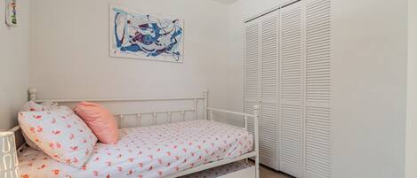 Uma cama branca com travesseiro rosa e colchão branco com padrões vermelhos, um criado-mudo branco com abajur, uma parede branca com um quadro e um armário branco com portas venezianas.
