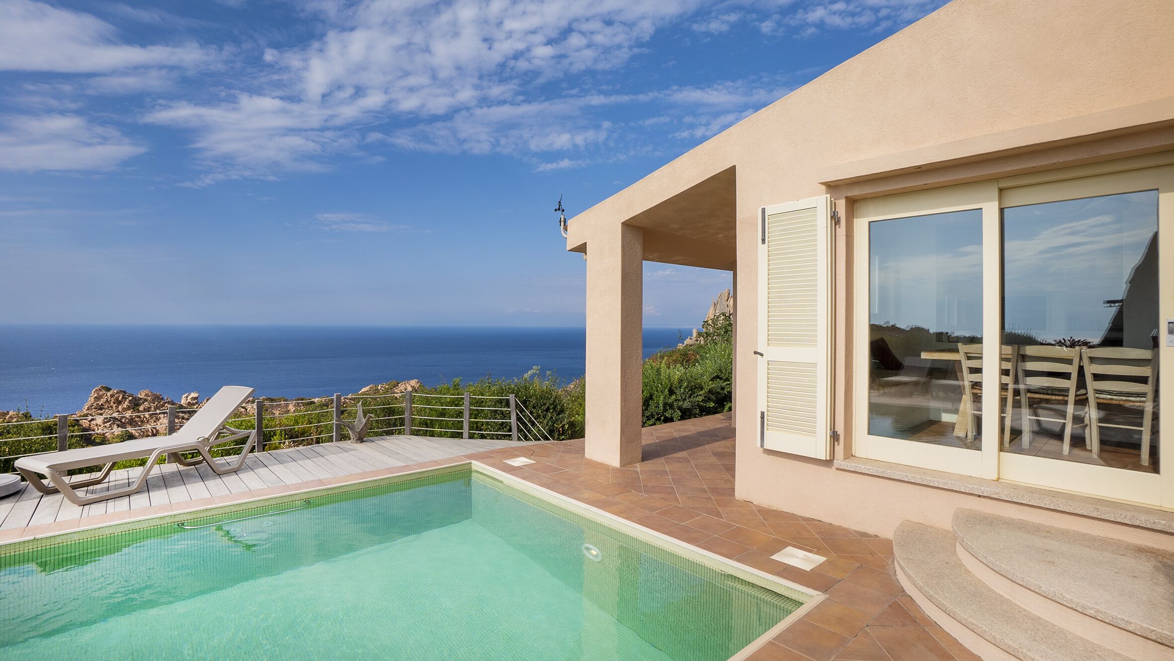 A swimming pool with a view of the ocean, a covered patio with chairs, and a large sliding glass door.