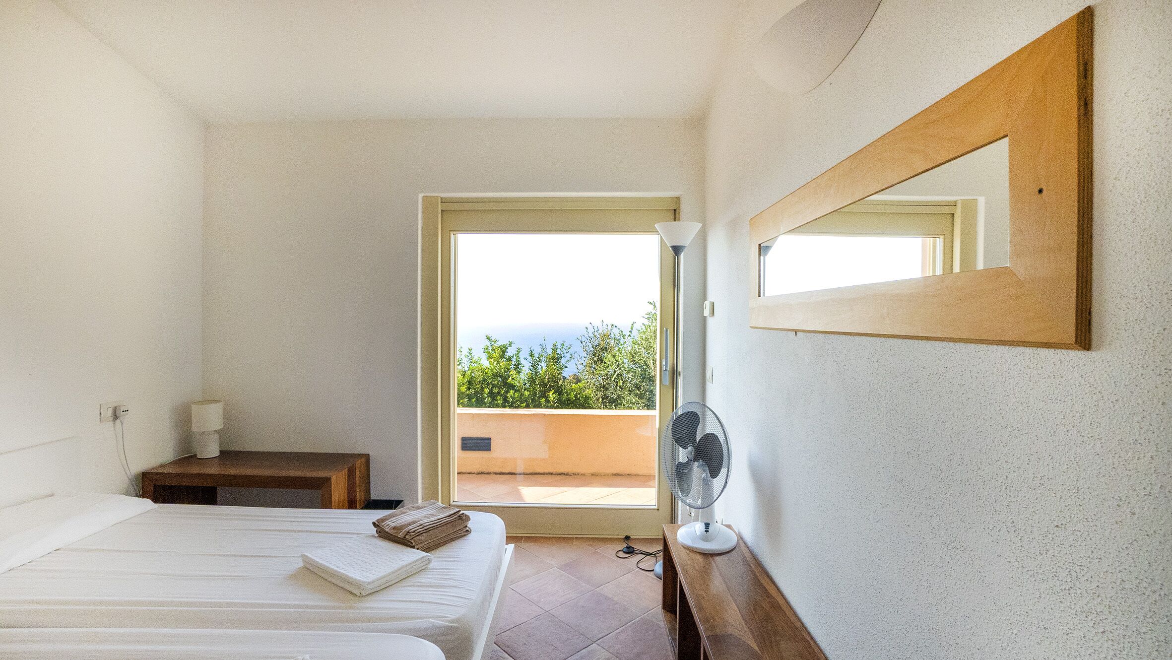 A bedroom with two beds, a bedside table, a mirror, a fan, and a view of the outdoors.