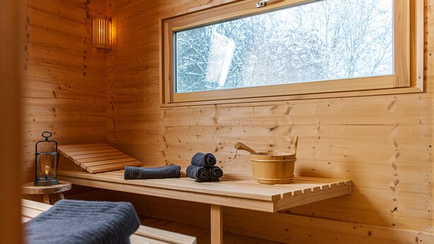 Interieur van een houten hutje met een bank, een tafel met handdoeken, een lantaarn en een raam met uitzicht op sneeuw.