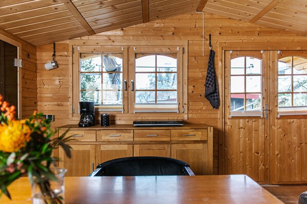A wooden interior with a kitchen area featuring a coffee maker, a small table, and a chair. There are two windows with curtains, and a towel hanging on the wall.