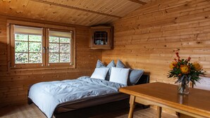 Een slaapkamer in een houten hutje met een bed, een klein tafeltje en een raam met jaloezieën.