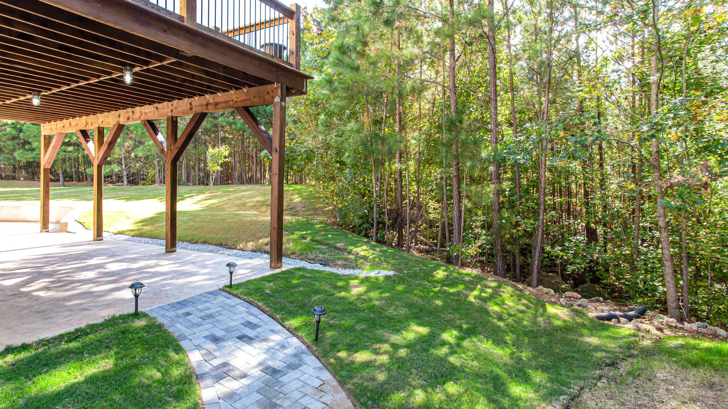 A covered outdoor area with a wooden pergola, a paved walkway, and a grassy lawn surrounded by trees.